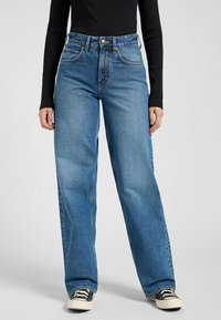 Hoch taillierte blaue Jeans mit lockerer Passform, ausgestattet mit einem Knopfverschluss, fünf Taschen und kontrastierenden Nähten, kombiniert mit einem schwarzen Oberteil und Sneakers.