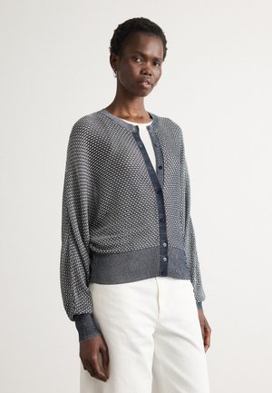 Kvinne iført en svart og hvit mønstret cardigan med ballongsleeves og mørke knapper, kombinert med hvite høytgående bukser.