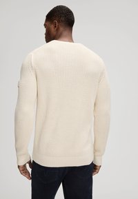 Kremowy sweter dzianinowy o teksturowanym, ażurowym wzorze. Posiada ściągacze na mankietach i u dołu. Na plecach widoczne są detale przeszyć.