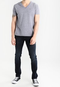 Man wearing a gray V-neck T-shirt, dark skinny jeans, black and white casual sneakers, standing against a plain white background.