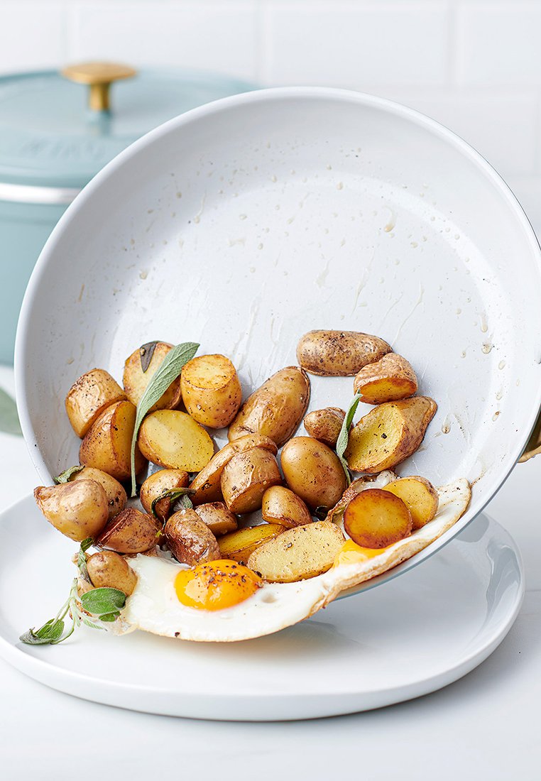 Plato de cerámica blanca con papas asadas dorado-marrón, un huevo frito y hierbas frescas, en ángulo mientras la comida se derrama de una sartén.