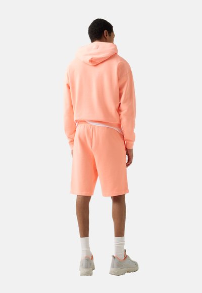 Mand iført en ferskenfarvet hættetrøje og matchende shorts, hvide sokker og lysegrå sneakers, stående med ryggen mod kameraet på en ensfarvet baggrund.