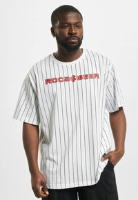 T-shirt oversize à rayures blanches avec logo rouge "ROCA" et "WEAR". Matière en coton, manches courtes, coupe décontractée, design à épaules tombantes.