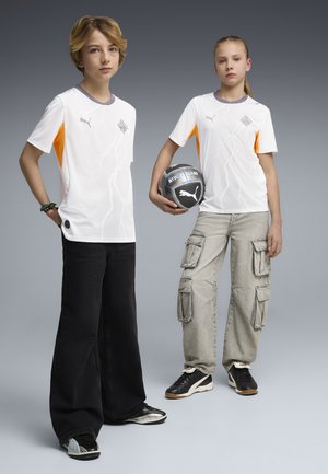 Deux jeunes portant des maillots de sport blancs se tiennent devant un fond gris, l'un tenant un ballon de football noir et blanc de la marque Puma.