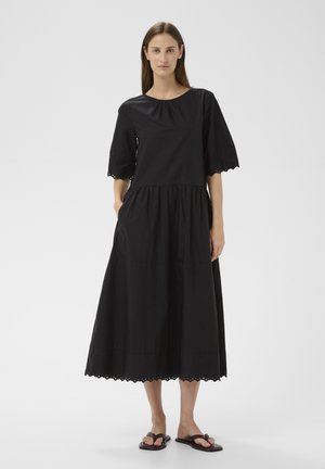 Moteris juoda midi suknele su bangelėmis apvadu ir pusilgomis rankovėmis, dėvinti juodus sandalus, ant balto vienodo fono.