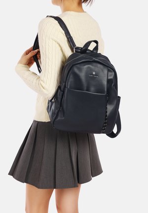 Zaino in pelle nera con una texture liscia, dotato di una tasca frontale, cerniere e spallacci regolabili, indossato sopra un maglione color crema e una gonna grigia.