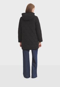 Manteau matelassé noir avec capuche, présentant un motif géométrique texturé. Le manteau est de longueur mi-longue, associé à un jean bleu foncé à jambe large.