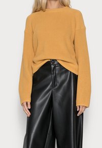 Sweter w kolorze musztardowej żółci z prążkowanym wzorem, o luźnym kroju i oversize'owych rękawach, zestawiony z wysokim stanem czarnych spodni z ekologicznej skóry.