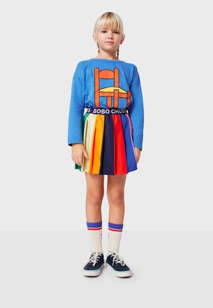 Ragazzina con trecce bionde che indossa una maglietta a maniche lunghe blu con una grafica di sedia arancione e una gonna plissettata a strisce arcobaleno.