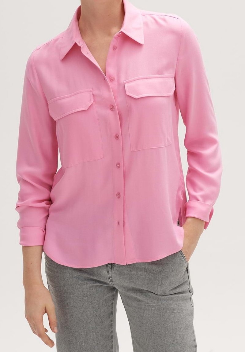 Femme portant une chemise rose à manches longues avec des poches poitrine à rabat et un jean gris, debout avec une main dans la poche.