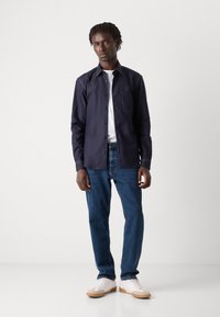 Navy shirt with button front and single chest pocket over a white t-shirt, paired with blue jeans and white sneakers with gum soles.