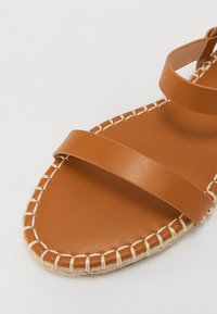 Sandal din piele maro, cu două curele late, talpă plată și margine din iută împletită. Textură netedă cu un design minim.