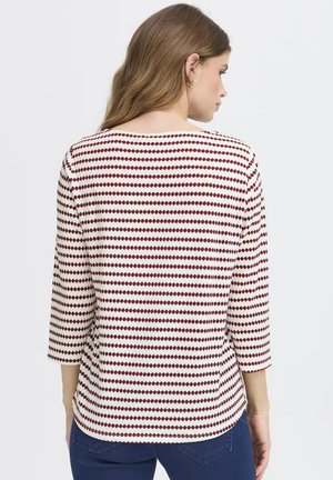 Donna con capelli lunghi che indossa un top bianco con righe orizzontali a punti color marrone scuro e maniche a tre quarti, di spalle.