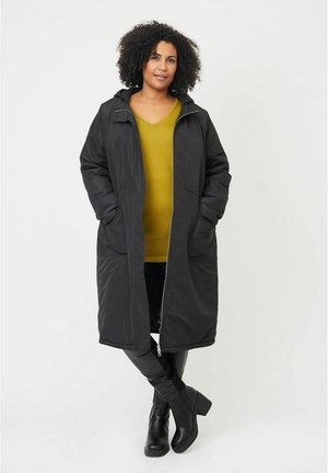 Schwarzer langer Mantel mit Reißverschluss vorne, Kapuze und zwei seitlichen Taschen, getragen über einem grünen V-Ausschnitt-Pullover. Das Model steht mit entspannten Armen und trägt kniehohe Stiefel.