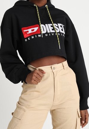 Henkilö pukeutuneena mustaan, lyhytmalliseen Diesel-huppariin, jossa on keltaiset kiristysnyörät, ja beigeihin, korkeavyötäröisiin cargo-housuihin, poseeraa yhdellä kädellä lantiolla.