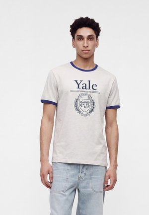 Jeune homme portant un t-shirt blanc de l’université Yale avec des bordures bleues et un jean bleu clair, debout devant un fond blanc uni.