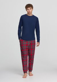 Maglietta a maniche lunghe blu marina con taschino sul petto abbinata a pantaloni del pigiama a scacchi rossi con accenti verdi e gialli, vestibilità ampia e polsini elastici.