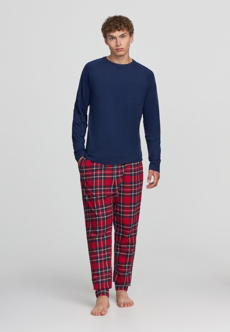 Maglietta a maniche lunghe blu marina con taschino sul petto abbinata a pantaloni del pigiama a scacchi rossi con accenti verdi e gialli, vestibilità ampia e polsini elastici.