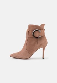 Bottines en suede couleur marron clair avec un bout pointu, une lanière enroulante et un accent métallique circulaire. Elles possèdent un talon aiguille.