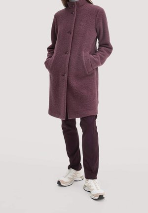 Kobieta w długim śliwkowym wełnianym płaszczu, ciemnych spodniach i białych trampkach, stojąca z rękami w kieszeniach na jednolitym tle.