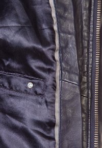 Intérieur d'une veste en cuir bleu marine montrant une doublure satinée lisse avec une petite poche et un bouton-pression, des coutures contrastées et une fermeture éclair.