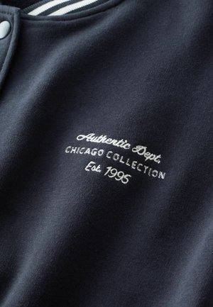 Jūrų pajėgų megztinis su baltais siuviniais: "Autentiška Dept. Chicago kolekcija, įkurta 1995", su tekstūrine medžiaga ir dryžuota apykakle.