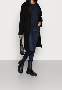 Long manteau noir, pull en maille torsadée bleu marine, jean foncé et bottines noires à semelles épaisses. Tient un petit sac à main noir. Textures lisses.