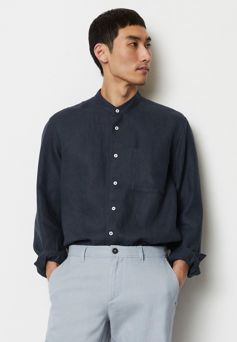 Marineblaues Leinenhemd mit Stehkragen, Brusttasche und weißen Knöpfen; kombiniert mit hellgrauen Hosen, die ein strukturiertes Muster aufweisen.