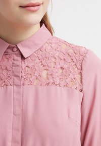 Lyserød bluse med knapkrave, prydet med blomsterret lace-detaljer på skuldrene og en blød, glat stofstruktur.
