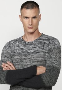 Pulover gri tricotat cu un model texturat. Are un guler rotund și mâneci negre în contrast. Logo mic pe piept.