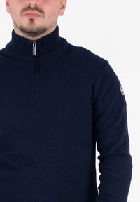 Mand iført en marineblå ribstrikket højhalset sweater med kvart lynlås og logo på det øverste venstre ærme, mod en ensartet baggrund.