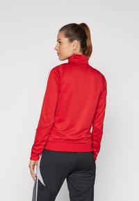 Red, long-sleeve athletic jacket with a high collar, featuring white stripe accents along the sleeves, made of smooth, stretchy fabric.