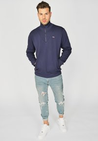 Sotapulloveri korkealla kauluksella ja puoleen vetoketjuun, valmistettu pehmeästä kankaasta. Yhdistetty kevyiden, kuluneiden farkkujen ja valkoisten tennareiden kanssa.