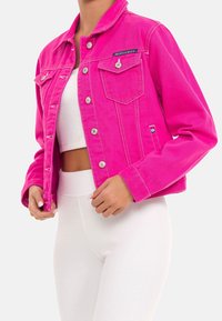 Lyserød denimjakke med krave, knaplukning foran, to brystlommer og kontrastsyninger. Båret over en hvid ribbet top og bukser.