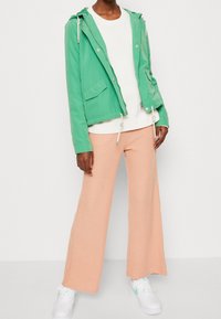 Zielona lekka kurtka z kapturem, przednimi kieszeniami, w zestawieniu z białym swetrem i brzoskwiniowymi szerokimi spodniami ze ściągaczem, dopełniony białymi sneakersami.