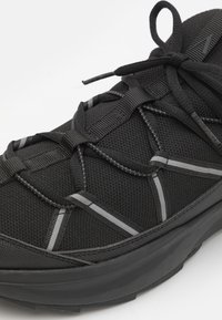 Svart atletisk sko med texturerad överdel, tjock sula, reflexdetaljer och korsade snören knutna i en rosett på en vit bakgrund.