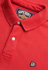 Polo rouge Superdry avec col plié, deux boutons blancs et logo brodé près du coin inférieur droit de l'image.