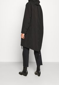 Person klädd i en lång svart kappa med huva, korta svarta byxor och svarta ankelstövlar, står med ryggen vänd.