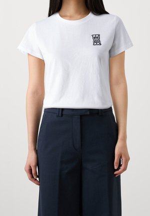 Camiseta blanca de algodón con mangas cortas y cuello redondo, con un pequeño logo negro bordado en el pecho izquierdo.