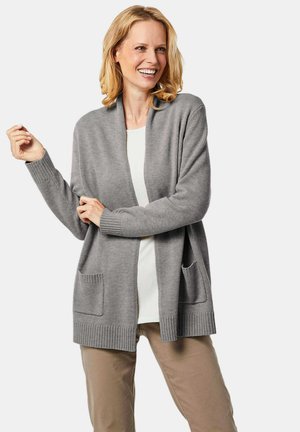 Grijze cardigan met open voorkant, geribde manchetten en zoom, met twee voorzakken. Draagt over een witte top met beige broek. Zachte textuur.