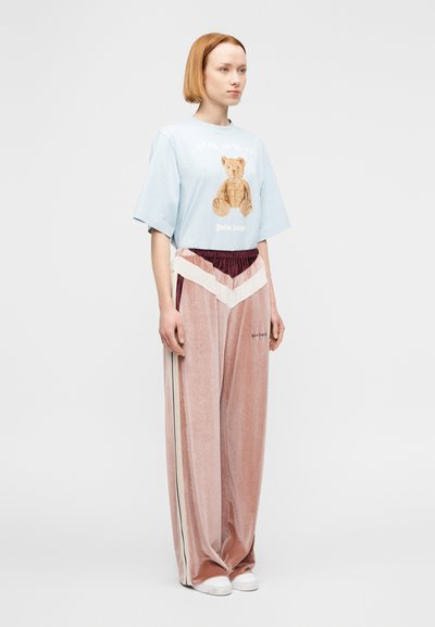 Γυναίκα με κοντά κόκκινα μαλλιά φοράει ανοιχτό μπλε μπλουζάκι με στάμπα αρκουδάκι και φαρδιά ρόζ παντελόνια βελούδου με λευκές ρίγες σε σχήμα ζικ-ζακ.