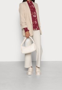 Blazer beige su una blusa floreale bordeaux, abbinato a pantaloni a zampa d'elefante crema e una borsa bianca intrecciata. Mocassini chunky beige completano il look.