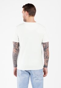 Man met full-arm tatoeages in een eenvoudige witte t-shirt en lichtblauwe jeans, staand met zijn rug naar ons toe tegen een witte achtergrond.