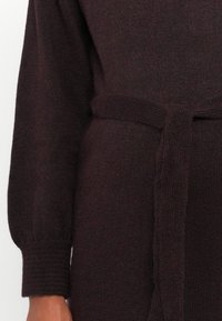Pletený sveter v tmavej burgundskej farbe s jemnou textúrou, ktorý má stiahnutý pás a rebrované manžety na rukávoch pre priliehavý vzhľad.