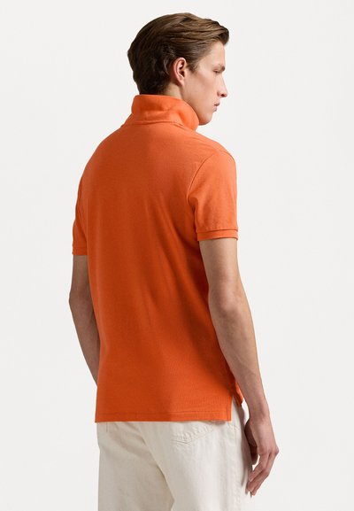 Orange polo-shirt i texturerad tyg, med krage och korta ärmar. Sömmen är något rundad med sidofrakar för ökad rörelsefrihet.