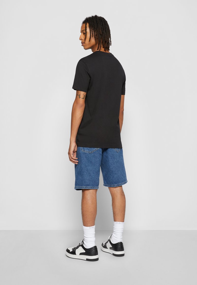 Joven con rastas que lleva una camiseta negra, pantalones cortos vaqueros azules, calcetines blancos y zapatillas negras y blancas, de pie de perfil.