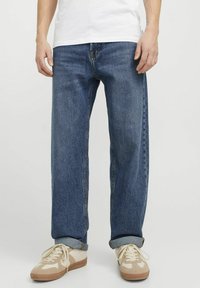 Jean en denim bleu ample avec des ourlets roulés, présentant un design classique à 5 poches et des coutures contrastantes. Portés avec des baskets beiges.