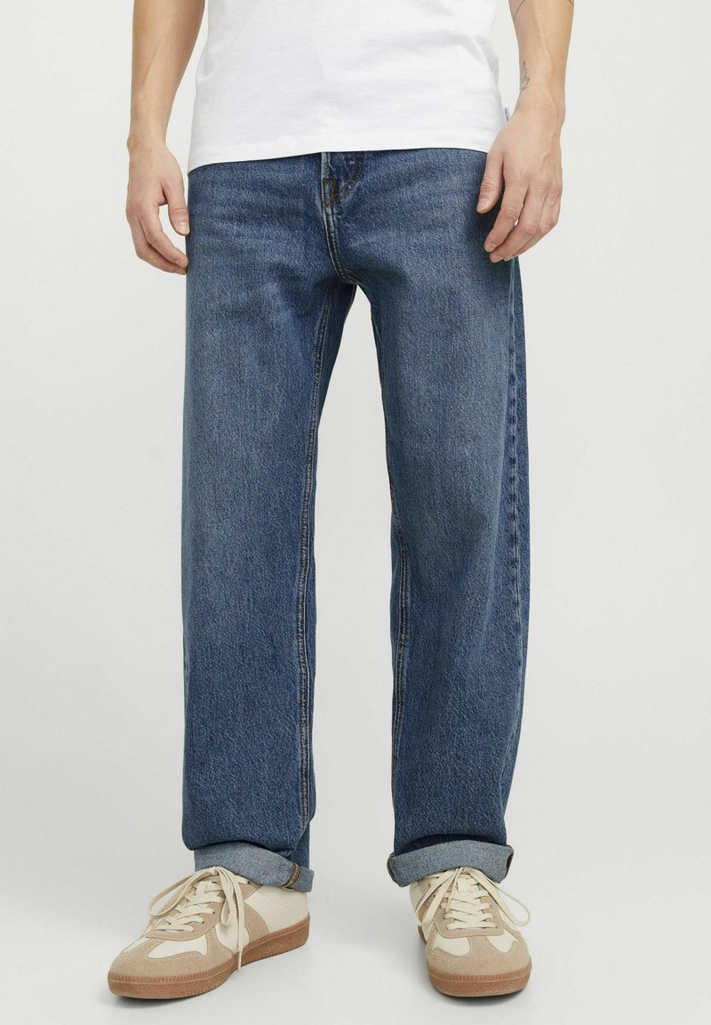 Jean en denim bleu ample avec des ourlets roulés, présentant un design classique à 5 poches et des coutures contrastantes. Portés avec des baskets beiges.
