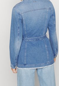 Chaqueta de mezclilla en azul claro con un cinturón en la cintura, que presenta mangas largas, un cuello y detalles de dobladillo deshilachado. La vista trasera muestra las líneas de las costuras.