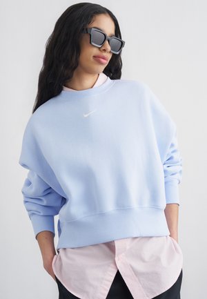 Kobieta z długimi ciemnymi włosami, w czarnych okularach przeciwsłonecznych, jasnoniebieskiej bluzie Nike na różowej koszuli zapinanej na guziki, ręce w kieszeniach.